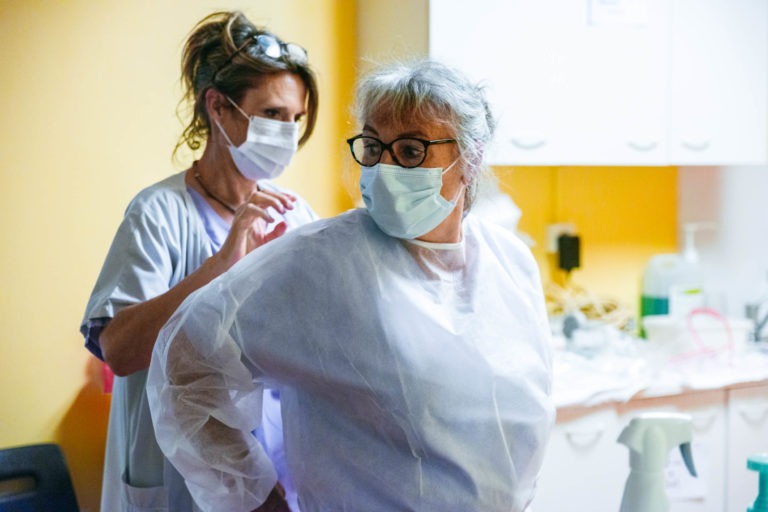 Un soignant aide une femme âgée à enfiler une blouse médicale blanche dans une clinique. Tous deux portent un masque de protection et la femme porte des lunettes. L'arrière-plan présente un mur jaune et du matériel médical sur un comptoir, soulignant le dévouement des Héros Méconnus du CH d'Angoulême.