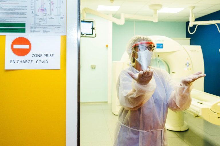 Un soignant en tenue de protection complète, comprenant un écran facial, un masque, une blouse et des gants, se tient dans une chambre d'hôpital à côté d'une porte jaune sur laquelle est écrit « Entrée interdite » et « Zone Prise En Charge COVID ». Du matériel médical et un scanner sont visibles en arrière-plan, véritable Héros Méconnus du CH d'Angoulême.