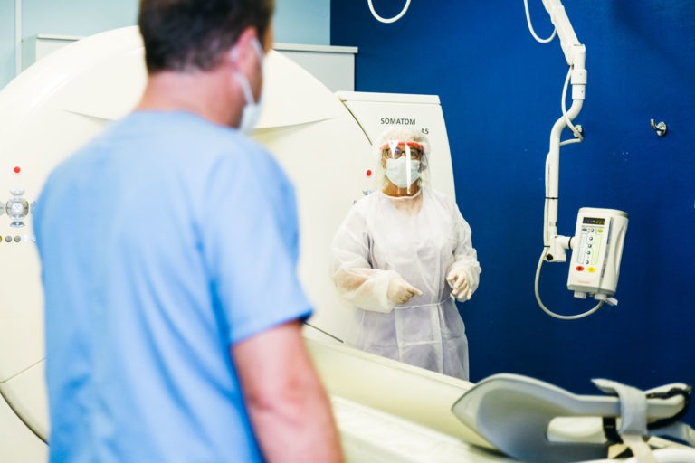 Un soignant en tenue de protection complète, comprenant un masque, une visière et une blouse, se tient à côté d'un scanner. Un patient portant un masque, vêtu d'une chemise bleue, se tient face au scanner. Le décor semble être une salle d'imagerie médicale avec des murs bleus et du matériel médical. Vraiment les Héros Méconnus du CH d'Angoulême au travail.