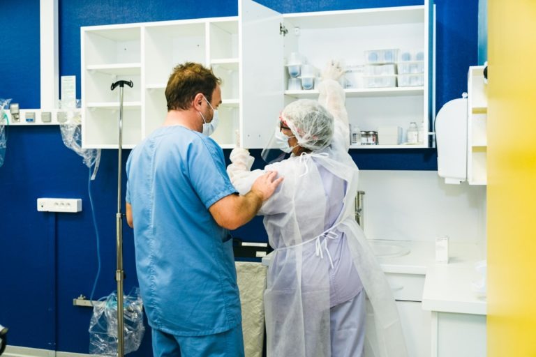 Deux professionnels de la santé dans une chambre d'hôpital. L'un, vêtu d'une blouse bleue et d'un masque chirurgical, aide un autre, en tenue de protection complète et avec un filet à cheveux, à accéder à une armoire suspendue. Du matériel et des fournitures médicales sont visibles sur les comptoirs et les étagères, avec des supports à perfusion en arrière-plan - de véritables Héros Méconnus du CH d'Angoulême.