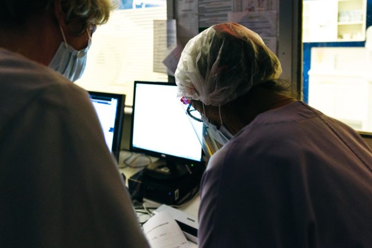 Deux agents de santé, les Héros Méconnus du CH d'Angoulême, portant des masques chirurgicaux et des charlottes de protection, travaillent dans une pièce faiblement éclairée. L'un se concentre sur des documents tandis que l'autre regarde un écran d'ordinateur. En arrière-plan, un autre moniteur et des papiers éparpillés.