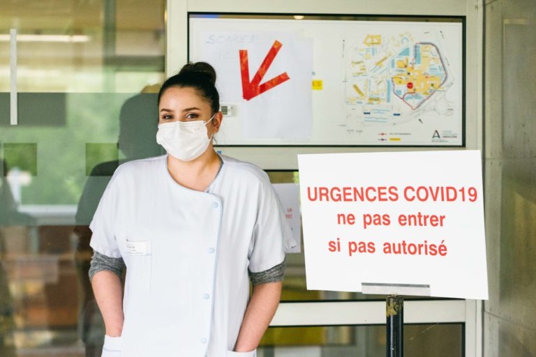 Une professionnelle de la santé en uniforme blanc et masque se tient à côté d'un panneau sur lequel est écrit en lettres rouges : « URGENCES COVID19 ne pas entrer si pas autorisé ». Derrière elle se trouve une porte vitrée avec une carte et une flèche rouge pointant vers la gauche, indiquant les directions à l'intérieur du bâtiment - de véritables Héros Méconnus du CH d'Angoulême.