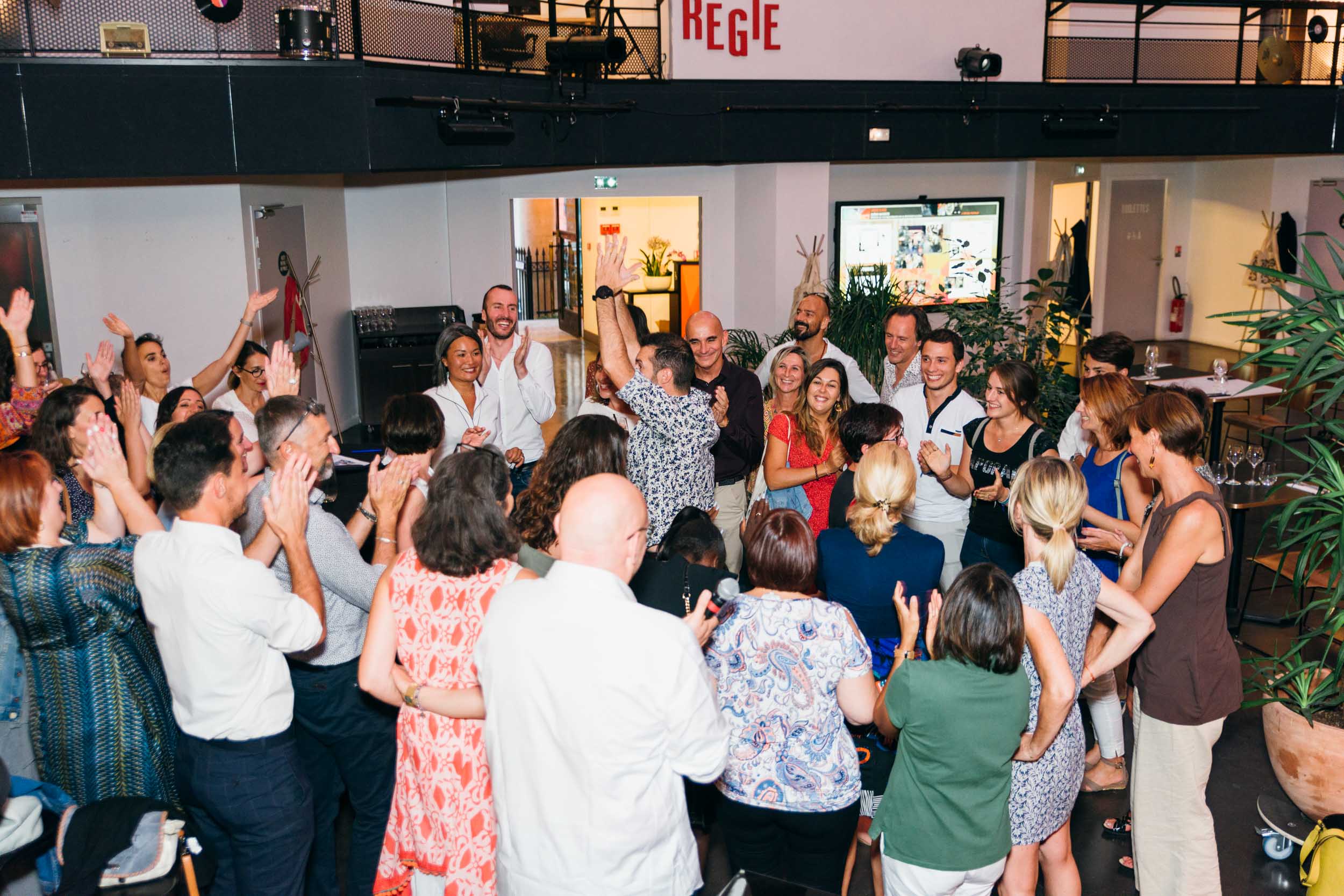 Un groupe de personnes applaudissent et acclament avec enthousiasme dans une salle lumineuse et moderne. Le groupe forme un demi-cercle autour d'un individu central levant la main en signe de célébration. La salle est ornée de plantes et de meubles modernes, avec une pancarte au-dessus de la porte sur laquelle est écrit "RÉGIE". Il s'agit d'un événement afterwork organisé par Ma Boîte d'Allumettes Bordeaux.