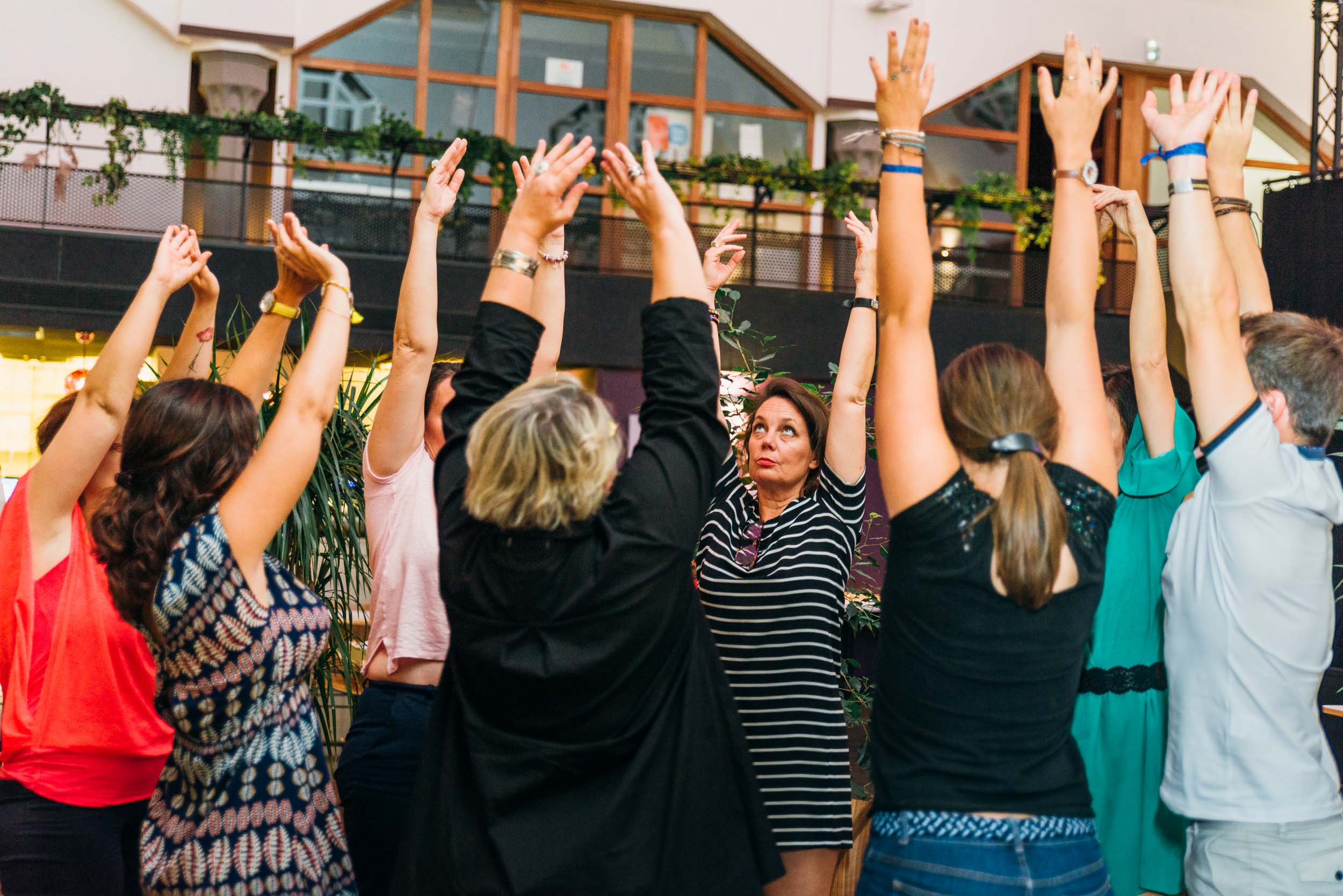 Un groupe de personnes, hommes et femmes, se tiennent en cercle à l'intérieur, les bras levés, les paumes tournées l'une vers l'autre. Engagées dans une joyeuse activité après le travail, certaines sont habillées de façon décontractée. De grandes fenêtres et de la verdure visible en arrière-plan ajoutent à l'ambiance animée de Ma Boîte d'Allumettes.