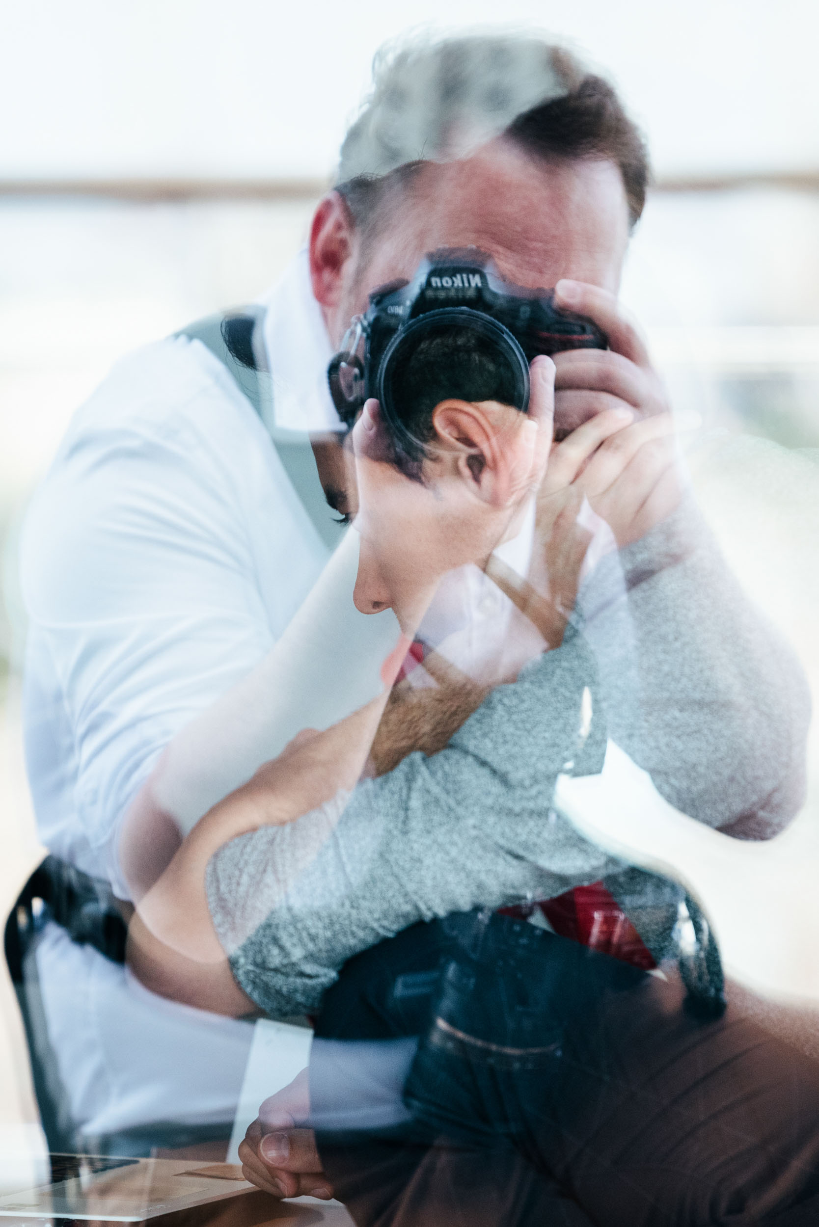 Un homme en chemise blanche tient un appareil photo Nikon devant ses yeux et prend une photo. L'image, parfaite pour un photographe d'entreprise, présente une superposition réfléchissante montrant une deuxième image partielle de lui et d'une autre personne, créant un effet de double exposition artistique. L'arrière-plan est légèrement flou, mettant en valeur les sujets principaux.