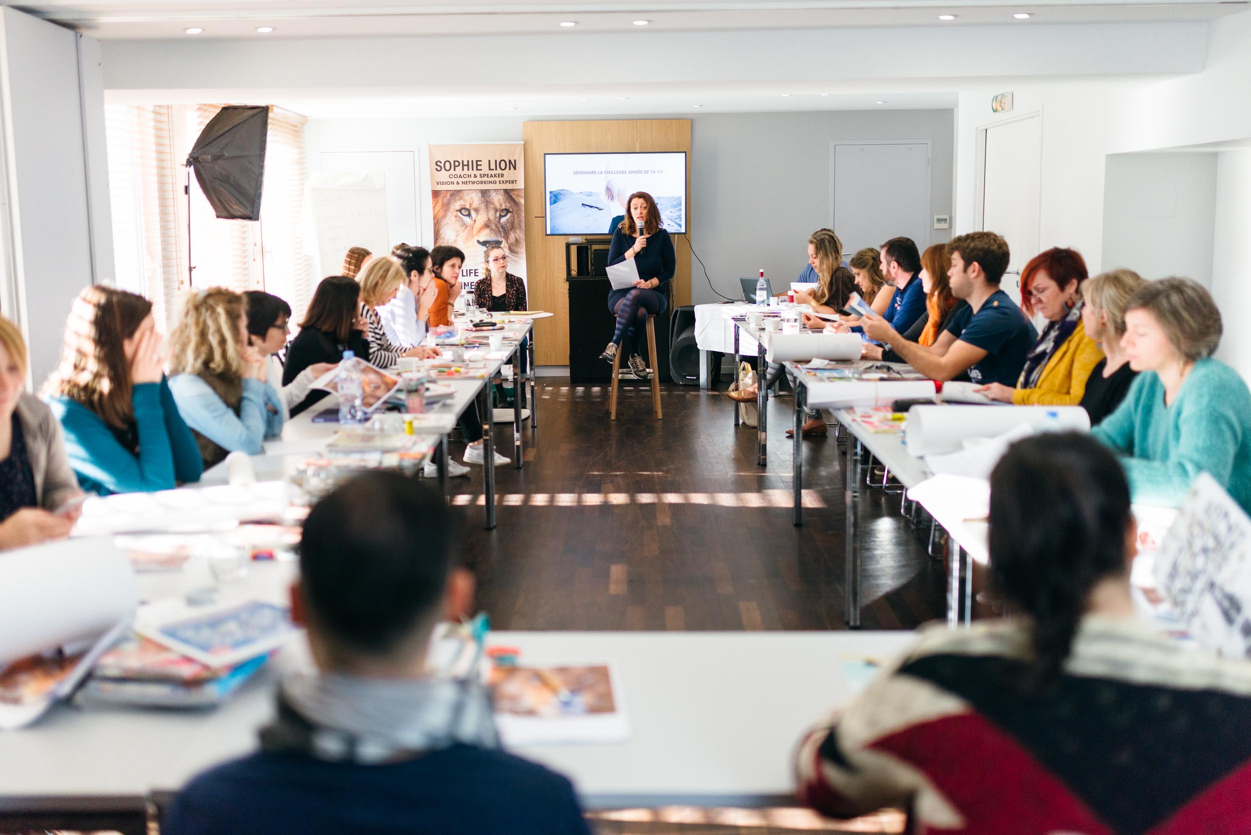 Une femme assise sur un tabouret au fond d'une salle lumineuse s'adresse à un groupe de participants au séminaire « La plus belle année de ta vie ». Des papiers à la main, elle parle pendant qu'ils prennent des notes. Un écran et des affiches, dont une mettant en scène Sophie LION, sont visibles derrière elle.