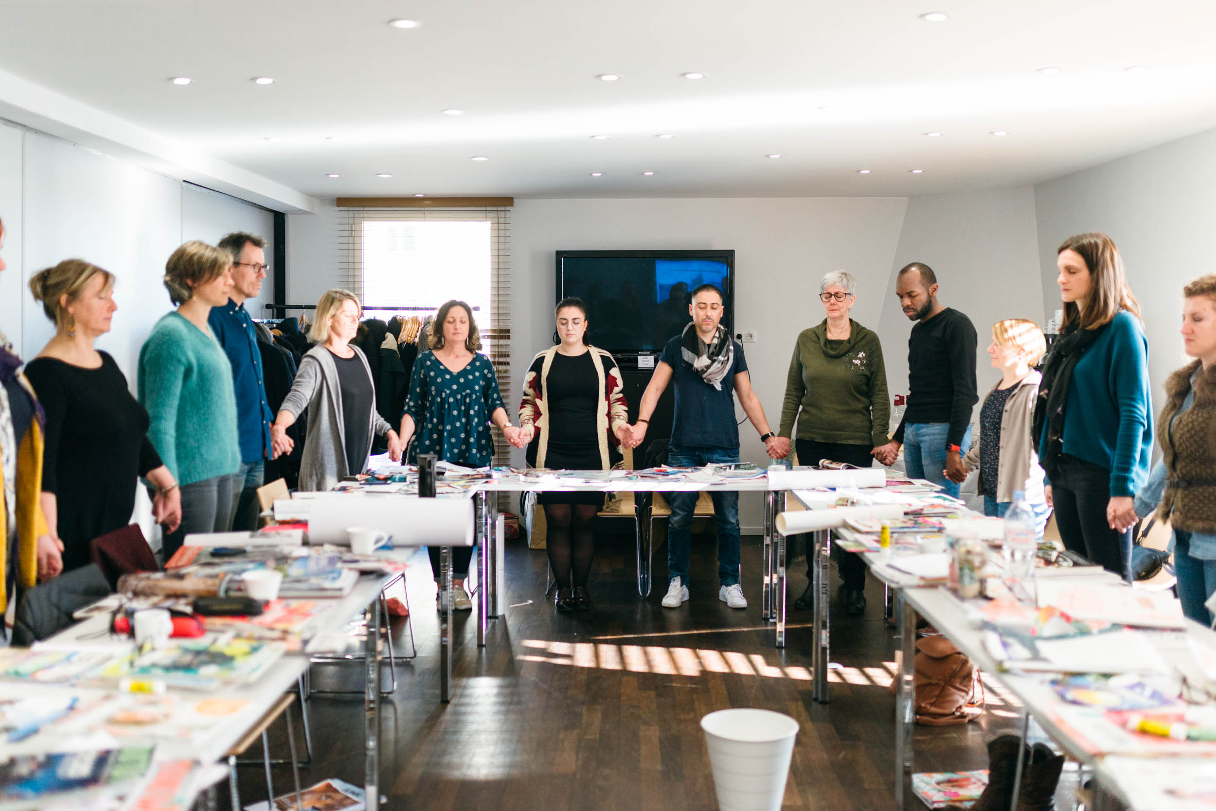 Un groupe hétéroclite de personnes se tient en cercle, main dans la main, dans une pièce bien éclairée avec une télévision au mur et des tables remplies de fournitures artistiques et de coupures de presse. Ils semblent unis et concentrés, participant probablement au séminaire « La plus belle année de ta vie » de Sophie LION. De grandes fenêtres laissent entrer la lumière naturelle.