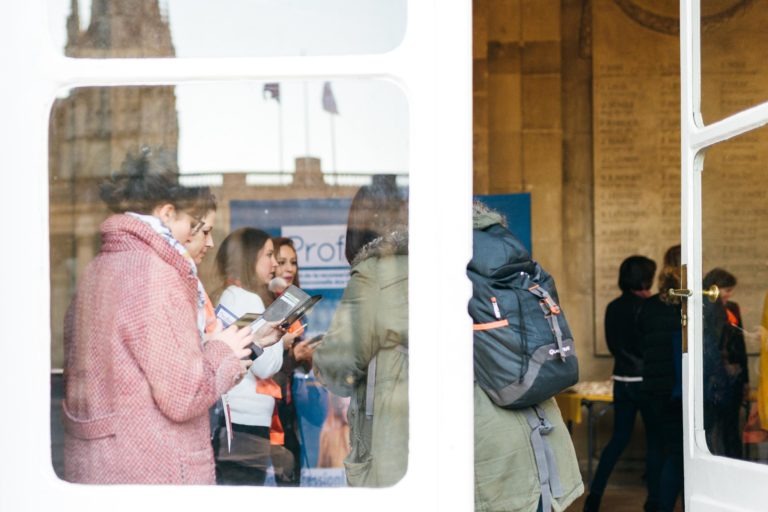 Un groupe de personnes est rassemblé à l'intérieur d'un bâtiment, vu à travers une grande fenêtre. Certains semblent en train de discuter tandis que d'autres regardent leur téléphone ou leur tablette. Des sacs à dos et des vestes suggèrent qu'ils se préparent pour le Salon Profession'L Bordeaux. Une bannière partiellement visible est en arrière-plan.