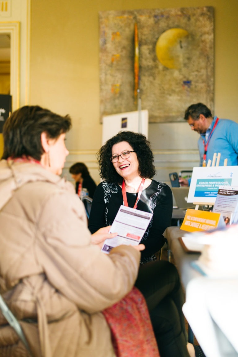 Une femme aux cheveux bouclés et portant des lunettes est assise à une table, souriante et tenant des documents. Elle discute avec une autre personne portant un manteau. En arrière-plan, un homme du Salon Profession'L Bordeaux lit quelque chose à une table sur laquelle sont exposés divers documents d'information. Un grand tableau abstrait décore le mur.