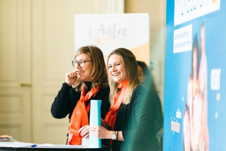 Deux femmes se tiennent devant un stand de conférence du Salon Profession'L Bordeaux, souriantes. Toutes deux portent des lunettes et ont les cheveux blonds détachés. Elles sont habillées en noir avec un foulard orange vif. Une femme tient un document et fait un geste de la main. L'arrière-plan présente une bannière floue et un autre affichage avec un texte indistinct.