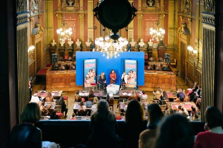 Un panel de quatre personnes est assis sur une scène dans une grande salle décorée de hauts plafonds, de lustres et de boiseries complexes. Un public regarde depuis le rez-de-chaussée et le balcon. Deux écrans bleus avec des publicités du Salon Profession'L Bordeaux sont placés derrière les panélistes. La salle est bien éclairée et élégante.