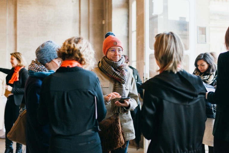 Un groupe de personnes vêtues de vêtements chauds, notamment d'écharpes, de chapeaux et de vestes, discutent dans un cadre intérieur bien éclairé. Certains tiennent des cahiers et discutent. L'ambiance est décontractée et amicale, un peu comme au Salon Profession'L Bordeaux. L'arrière-plan montre une grande fenêtre et des murs aux tons neutres.