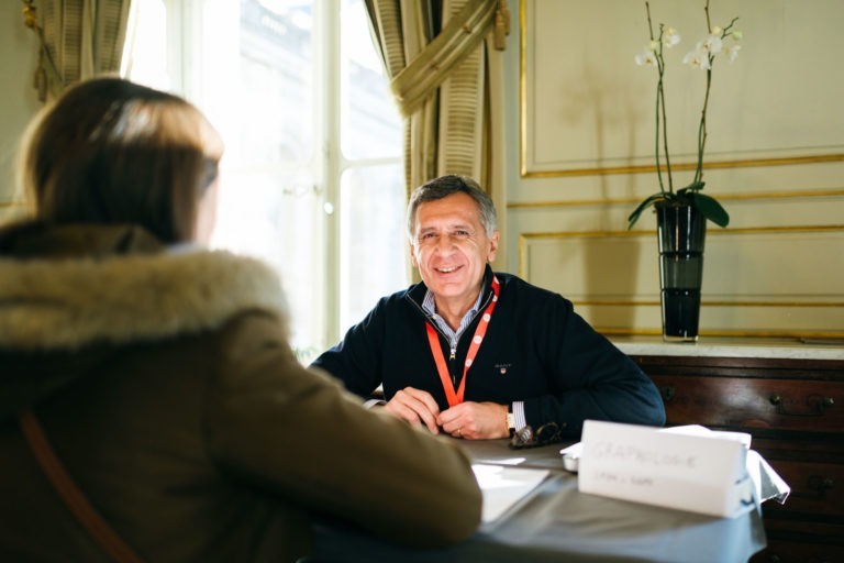 Un homme portant un pull noir et un cordon rouge autour du cou sourit, assis derrière une table sur laquelle est écrit « GRAPHCORE » au Salon Profession'L Bordeaux. Il parle à une personne aux cheveux longs et bruns, vêtue d'une veste verte avec une capuche doublée de fourrure, dans une pièce bien éclairée, ornée de rideaux élégants et d'une orchidée en pot.