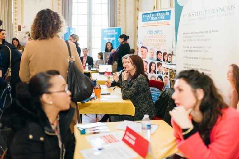 Le Salon Profession'L de Bordeaux, un salon de l'emploi très animé, propose des stands avec des représentants assis à des tables drapées de jaune, qui dialoguent avec les participants. Diverses bannières affichent du texte en français, faisant la promotion de programmes de formation et de coaching. Les gens sont animés, discutent des opportunités, avec en arrière-plan une salle lumineuse et élégante.