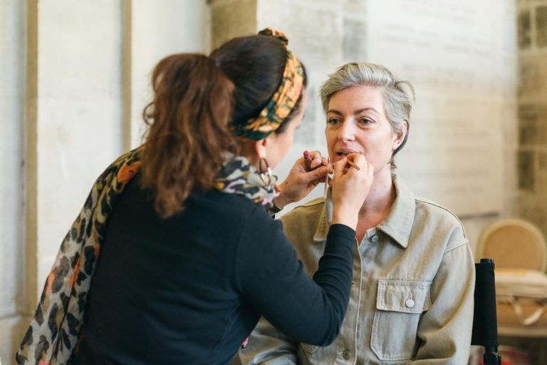 Une maquilleuse applique du rouge à lèvres sur une personne aux cheveux gris courts, assise. La maquilleuse, aux cheveux noirs et coiffée d'un bandeau et d'un foulard colorés, se concentre intensément sur son travail. Elles se trouvent dans le Salon Profession'L Bordeaux, une pièce aux murs clairs et à la lumière douce et naturelle.