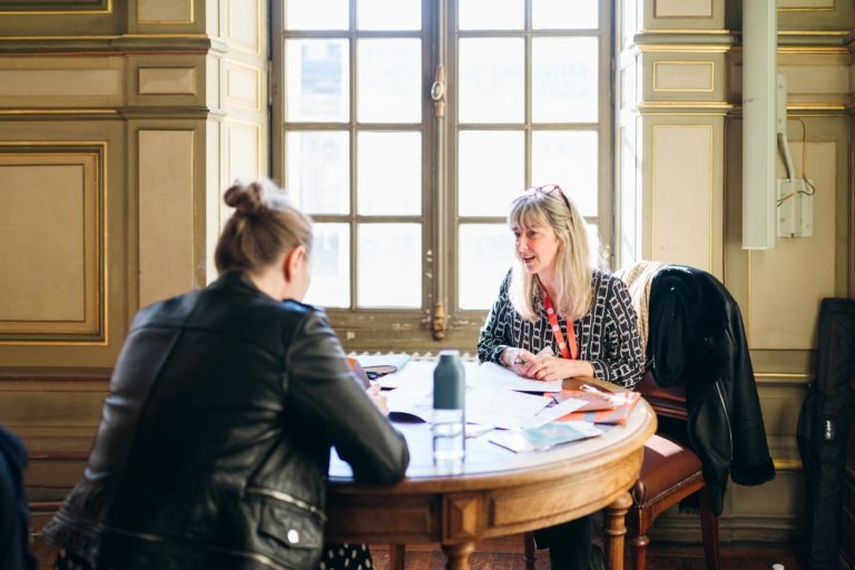 Dans le Salon Profession'L Bordeaux, une femme aux longs cheveux blonds, vêtue d'une chemise à motifs, est assise à une table ronde en bois recouverte de papiers et d'une bouteille d'eau. Elle discute avec une autre femme aux cheveux en chignon, vêtue d'une veste noire. Elles se trouvent dans une pièce ornée de grandes fenêtres et de murs décorés.