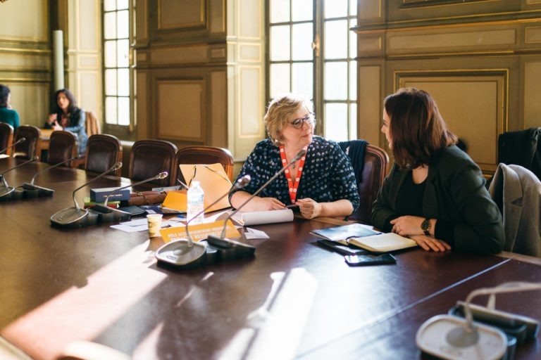 Deux femmes sont assises à une grande table de conférence dans une salle aux murs lambrissés et aux hautes fenêtres du Salon Profession'L Bordeaux. L'une porte un chemisier à pois bleu et un cordon rouge, et l'autre un blazer foncé. Elles discutent avec des papiers, une bouteille d'eau et des microphones devant elles. D'autres personnes sont floues en arrière-plan.