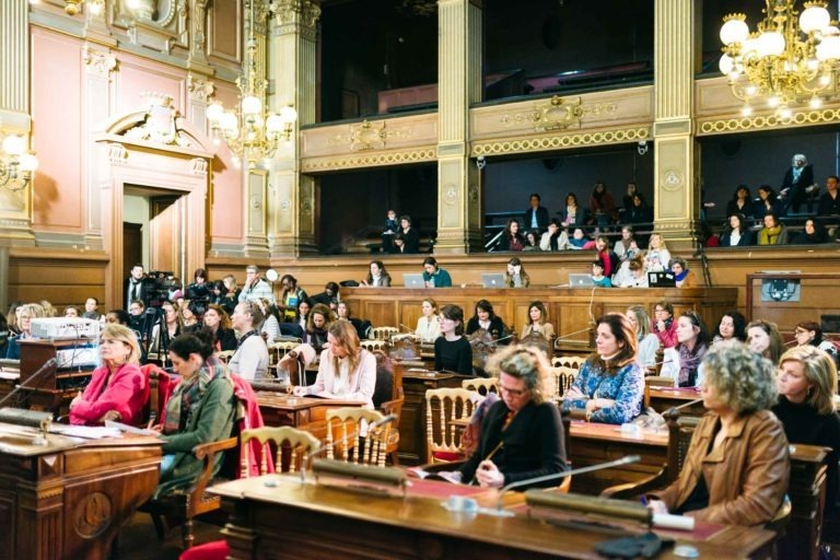 Un grand groupe de personnes, principalement des femmes, sont assises dans une salle historique et décorée avec de hauts plafonds et des lustres en or au Salon Profession'L Bordeaux. Le public est concentré sur un orateur qui n'est pas visible. Les murs sont ornés de boiseries complexes et il y a des individus dans la galerie au-dessus.