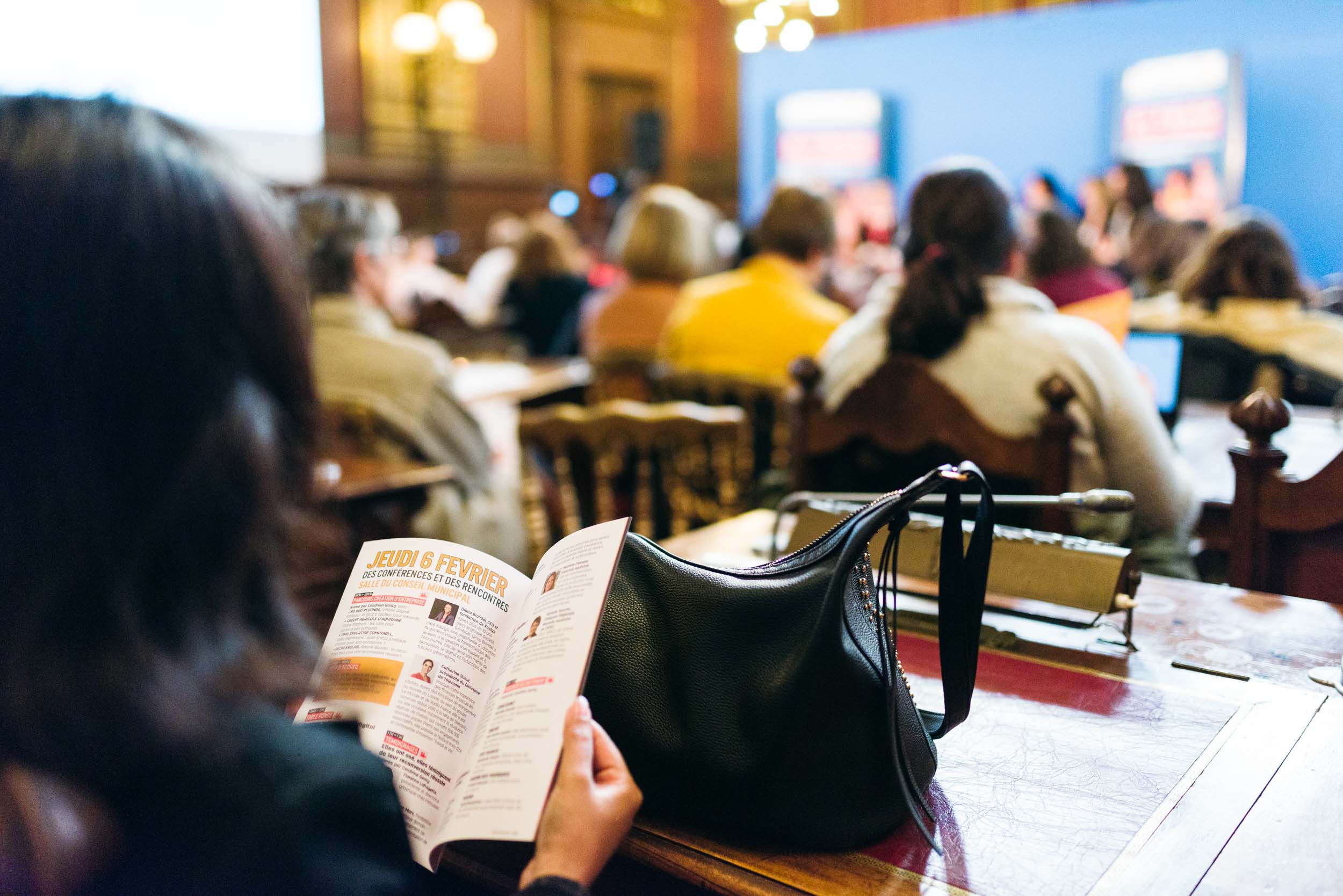 Des personnes sont assises dans une salle de conférence décorée de boiseries et de lustres. Une personne au premier plan, tenant une brochure du Salon Profession'L Bordeaux, est mise au point, tandis qu'un sac à main noir repose sur la table. L'arrière-plan montre une présentation sur un écran bleu et un public flou tourné vers l'avant.