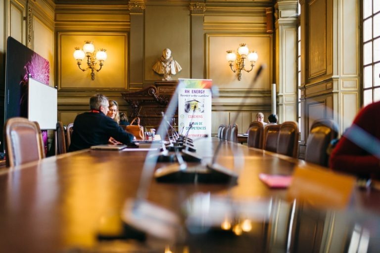 Une grande salle de conférence avec des lustres ouvragés et des boiseries évoque l'élégance du Salon Profession'L Bordeaux. Une longue table avec des microphones est installée au centre. Quelques personnes sont assises, avec une bannière en arrière-plan affichant "RH Energy". Un buste de statue est accroché au mur derrière eux. La lumière du soleil entre par de hautes fenêtres.