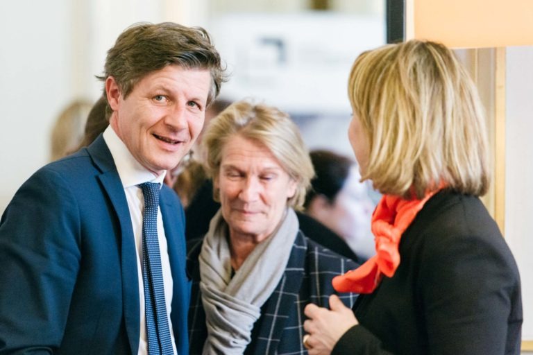 Trois personnes discutent dans une salle bien éclairée du Salon Profession'L Bordeaux. L'homme à gauche, vêtu d'un costume bleu et d'une cravate, regarde directement la caméra. Il se tient à côté d'une femme plus âgée portant une écharpe grise et un blazer à carreaux. À droite, une femme blonde porte un blazer noir et un foulard orange vif.