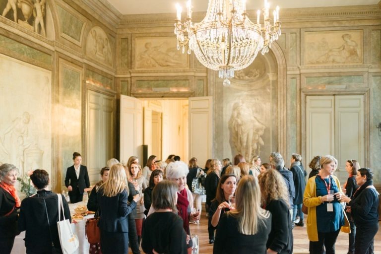 Un grand groupe de personnes est réuni dans une salle décorée avec de hauts plafonds, un grand lustre et des peintures murales classiques. Ils discutent et semblent assister à un événement du Salon Profession'L Bordeaux. Beaucoup tiennent des boissons et une table avec des rafraîchissements est visible sur la gauche.