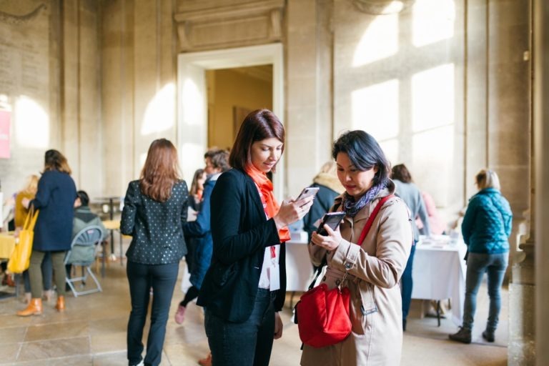 Deux femmes se tiennent au centre d'une pièce aux hauts plafonds, les yeux rivés sur leur téléphone. L'une porte un blazer noir et une écharpe rouge, l'autre un manteau beige et un sac à main rouge. Autour d'elles, au Salon Profession'L Bordeaux, les gens s'adonnent à diverses activités et la lumière du soleil pénètre à travers de grandes fenêtres, illuminant la scène.