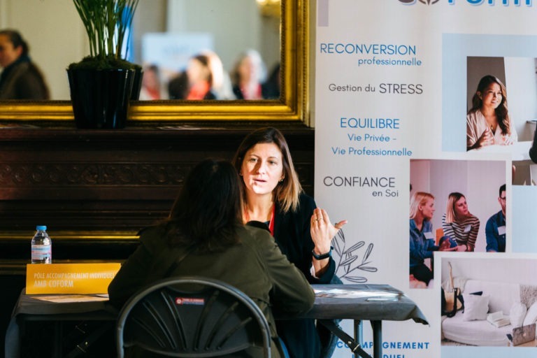 Deux femmes sont assises à une petite table et discutent. L'une d'elles regarde vers l'avant tout en faisant des gestes de la main, tandis que l'autre tourne le dos à la caméra. Une grande bannière derrière elles fait la promotion du Salon Profession'L Bordeaux avec un texte en français sur le coaching professionnel, la gestion du stress et le renforcement de la confiance en soi.