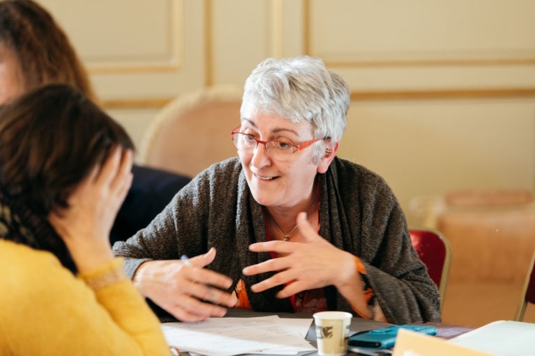 Une femme âgée aux cheveux gris courts et portant des lunettes parle avec animation tout en gesticulant avec ses mains au Salon Profession'L Bordeaux. Elle est assise à une table avec une autre personne, qui a la main sur le visage. Des papiers et une tasse de café sont devant eux, dans ce qui semble être un environnement intérieur décontracté.