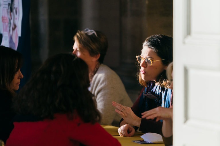 Un groupe de quatre personnes est assis autour d'une table jaune et discute au Salon Profession'L Bordeaux. Une femme portant des lunettes et un foulard bleu parle avec animation, en faisant des gestes avec ses mains. Deux autres personnes sont dans l'ombre partielle et une porte sur la droite est légèrement entrouverte, laissant entrer la lumière.