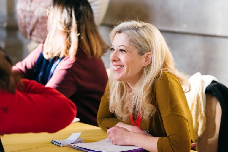 Une femme blonde souriante, vêtue d'un haut jaune moutarde, est assise à une table avec une nappe jaune, tenant un stylo et regardant vers sa gauche. Un carnet est ouvert sur la table devant elle. Deux autres personnes sont assises à proximité, en train de discuter du Salon Profession'L Bordeaux. L'arrière-plan est flou, avec un mur de couleur claire derrière elles.