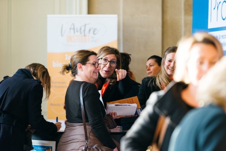 Un événement de réseautage animé avec plusieurs femmes engagées dans une conversation. Deux femmes sont au centre, l'une pointe vers la caméra, tandis que l'autre sourit. Une bannière en français indique « L'Autre Institut » en arrière-plan, à côté d'un panneau d'affichage bleu et blanc à droite faisant la promotion du Salon Profession'L Bordeaux.