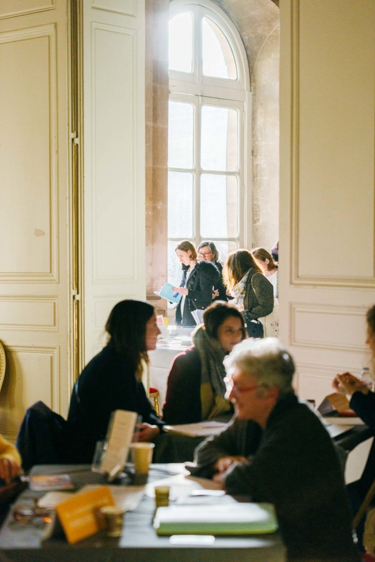 Dans une grande salle haute de plafond et dotée de grandes fenêtres, les gens discutent et s'amusent autour de tables. Certains sont assis et discutent, d'autres se tiennent debout en groupe, tenant des documents. La lumière chaude du soleil remplit le Salon Profession'L Bordeaux, créant une atmosphère accueillante et animée.