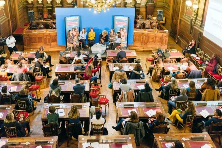 Un grand groupe de personnes est assis dans une salle décorée avec des bureaux et des chaises en bois, face à une scène sur fond bleu au Salon Profession'L Bordeaux. Sur la scène, un panel d'intervenants discute. Les participants prennent des notes et écoutent attentivement, avec un écran et des bannières visibles en arrière-plan.