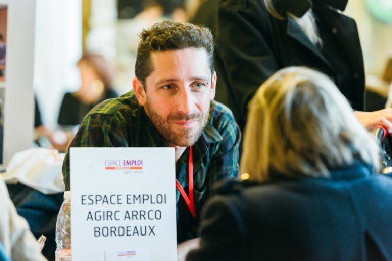 Un homme aux cheveux bruns courts et bouclés et portant une barbe est assis derrière une pancarte sur laquelle est écrit « ESPACE EMPLOI AGIRC ARRCO BORDEAUX » au Salon Profession'L Bordeaux. Il discute avec une femme blonde qui tourne le dos à la caméra. L'arrière-plan est flou, ce qui indique un environnement animé.