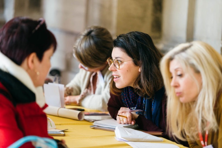 Quatre femmes sont assises à une table recouverte de papier jaune, en pleine conversation et en train de remplir des papiers au Salon Profession'L Bordeaux. La femme au centre, portant des lunettes et des cheveux noirs, se penche en avant avec attention. Deux autres femmes, l'une en manteau rouge et l'autre aux cheveux blonds, sont partiellement visibles de chaque côté d'elle.