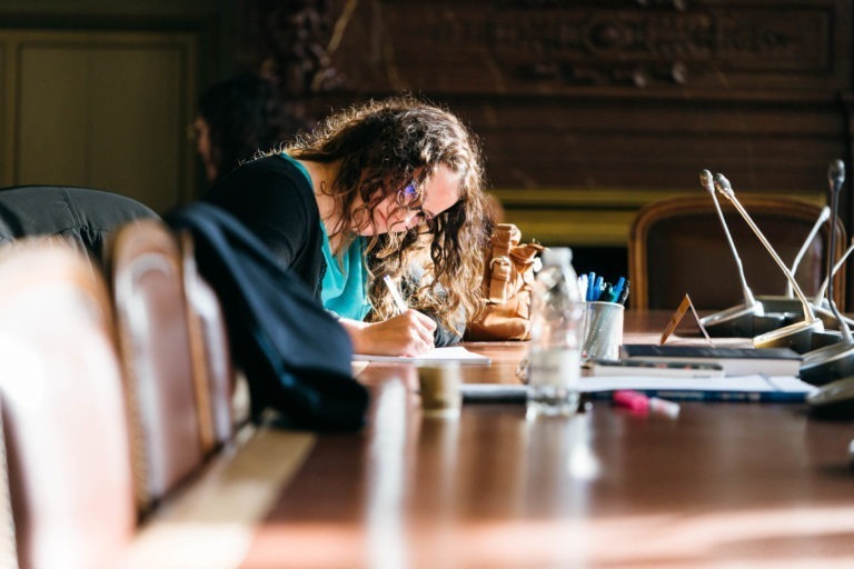 Une femme aux cheveux bouclés et aux lunettes est assise à une grande table en bois, penchée en avant et écrivant sur un morceau de papier. La table est encombrée de divers objets, dont des stylos, des bouteilles et des documents, créant une atmosphère rappelant le Salon Profession'L Bordeaux. Elle semble concentrée sur sa tâche dans une pièce bien éclairée avec des boiseries élaborées sur les murs.