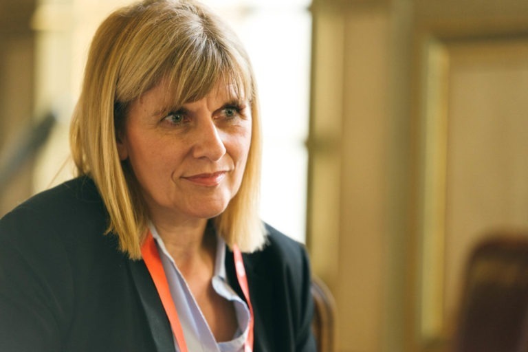 Une femme aux cheveux blonds mi-longs et à la frange, porte un blazer noir sur un chemisier bleu clair. Elle a un cordon rouge autour du cou et regarde légèrement vers la gauche avec une expression pensive, comme si elle envisageait sa prochaine visite au Salon Profession'L Bordeaux. L'arrière-plan est flou, suggérant un décor intérieur.