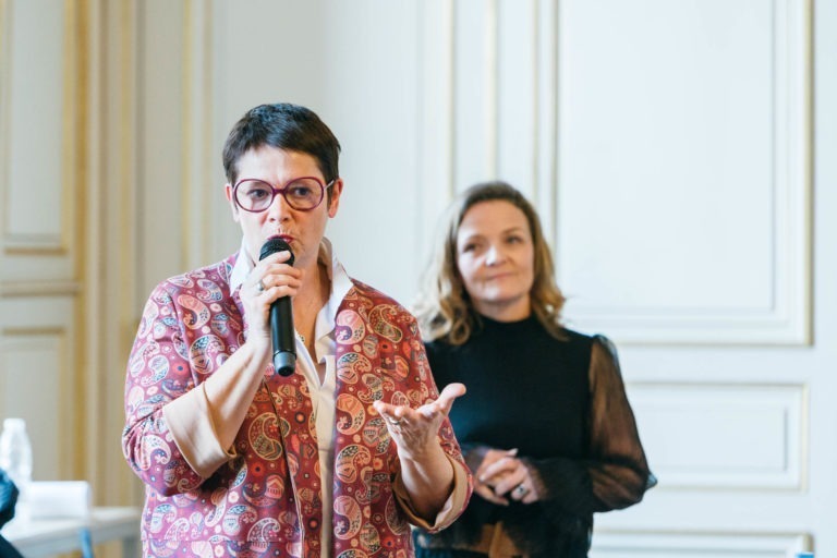 Une personne aux cheveux courts et aux grandes lunettes rondes, vêtue d'un blazer à motifs roses sur une chemise blanche, parle au micro au Salon Profession'L Bordeaux. Une autre personne aux cheveux mi-longs, vêtue d'une tenue noire, se tient en arrière-plan flou. Elles se trouvent dans une pièce aux boiseries décoratives de couleur crème.