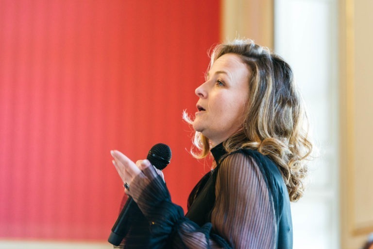 Une femme aux cheveux blonds ondulés parle dans un micro tenu dans sa main droite. Elle porte un haut à manches longues foncé avec des manches transparentes. L'arrière-plan présente un mur rouge texturé à gauche et un mur de couleur claire à droite, suggérant qu'elle pourrait être au Salon Profession'L Bordeaux en train de prononcer un discours ou une présentation.