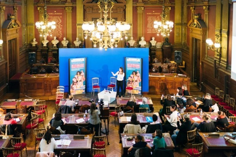 Une femme se tient debout sur une scène avec un fond bleu, présentant au Salon Profession'L Bordeaux dans une grande salle décorée de lustres et d'un décor classique. Les participants sont assis à des tables, face à la scène, écoutant attentivement. Deux affiches de chaque côté de la scène présentent des images et des textes similaires.