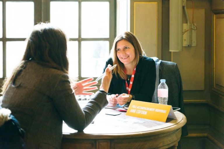 Deux femmes sont assises à une table ronde près d'une fenêtre. L'une, aux cheveux longs, tourne le dos et fait des gestes avec ses mains, tandis que l'autre, portant un cordon et souriant, regarde vers l'avant. Une pancarte jaune sur la table indique « Pôle accompagnement individuel AN COPAIN ». Une bouteille d'eau et une brochure du Salon Profession'L Bordeaux sont également posées sur la table.