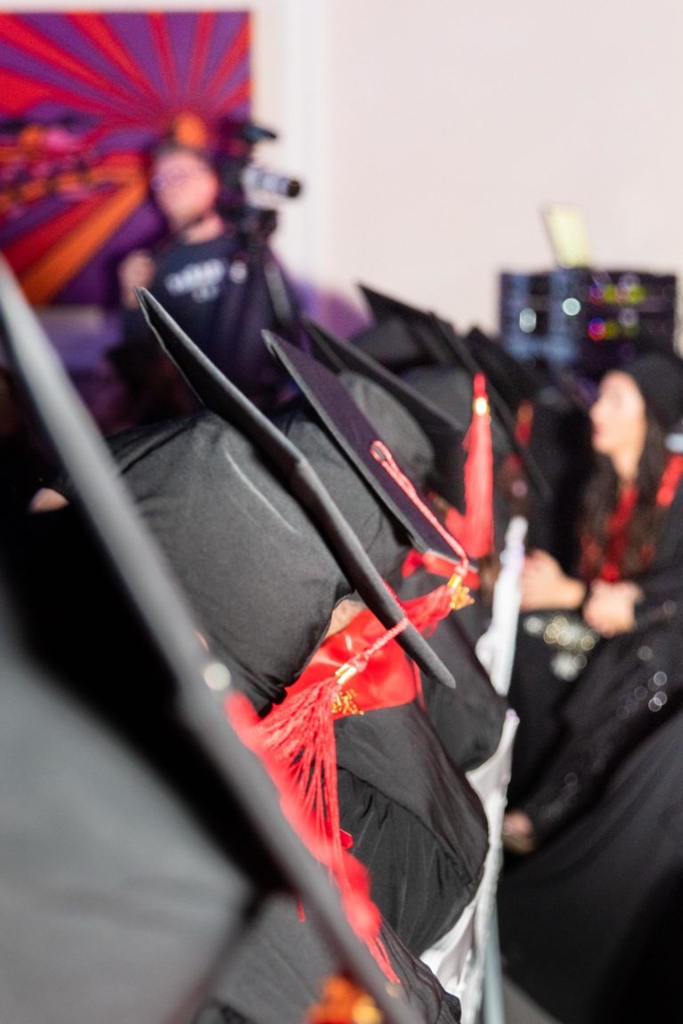 Une rangée de diplômés vêtus de toges noires et de casquettes académiques carrées est assise à l'intérieur d'une salle. Des glands rouges pendent de leurs casquettes, marquant la Cérémonie de remise des diplômes EBBS. En arrière-plan, une personne utilise une caméra et une autre personne vêtue de noir est assise. Une œuvre d'art colorée décore le mur.