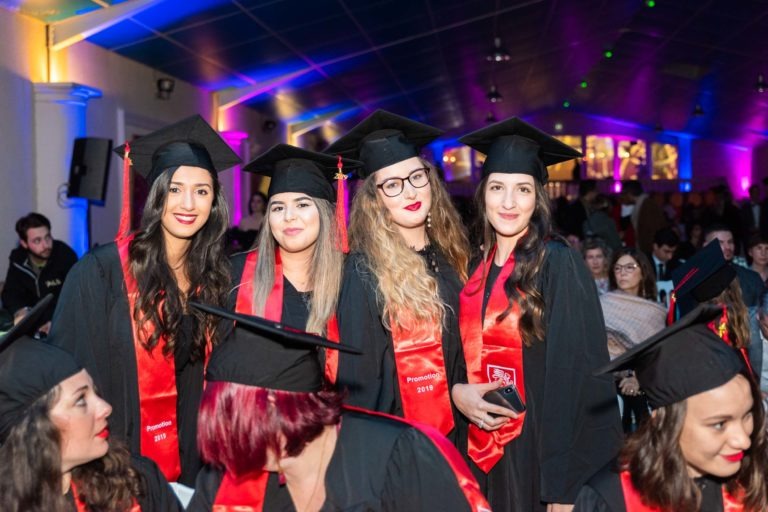 Un groupe de cinq diplômés souriants, portant des toges et des casquettes noires avec des écharpes rouges, se tiennent ensemble lors de la cérémonie de remise des diplômes de l'EBBS. Ils sont à l'intérieur, sous un plafond éclairé de bleu et de violet. Derrière eux se trouvent d'autres participants et diplômés. L'un des diplômés à droite tient un smartphone.