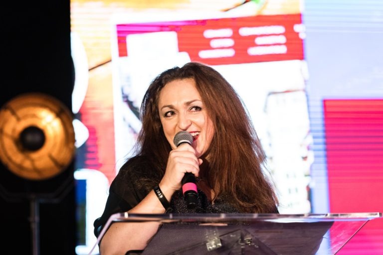 Une femme aux longs cheveux bruns se tient debout sur un podium bien dégagé, souriant et tenant un micro. Elle porte un haut noir et porte un bracelet au poignet gauche. Derrière elle, lors de la cérémonie de remise des diplômes de l'EBBS, se trouve un écran numérique coloré et flou affichant divers graphiques et textes, avec un projecteur rond à gauche.