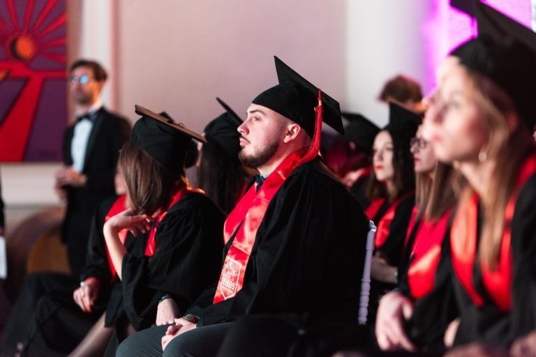 Un groupe de diplômés portant des toges et des casquettes noires avec des étoles rouges sont assis attentivement pendant leur cérémonie de remise des diplômes EBBS. Un diplômé au centre, bien visible, semble concentré. L'arrière-plan présente des œuvres d'art colorées et une personne en tenue de soirée debout. L'atmosphère est festive.