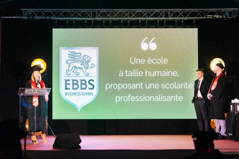 Une femme se tient sur une scène avec un podium et un écran représentant un lion, accueillant la Cérémonie remise diplômes EBBS.