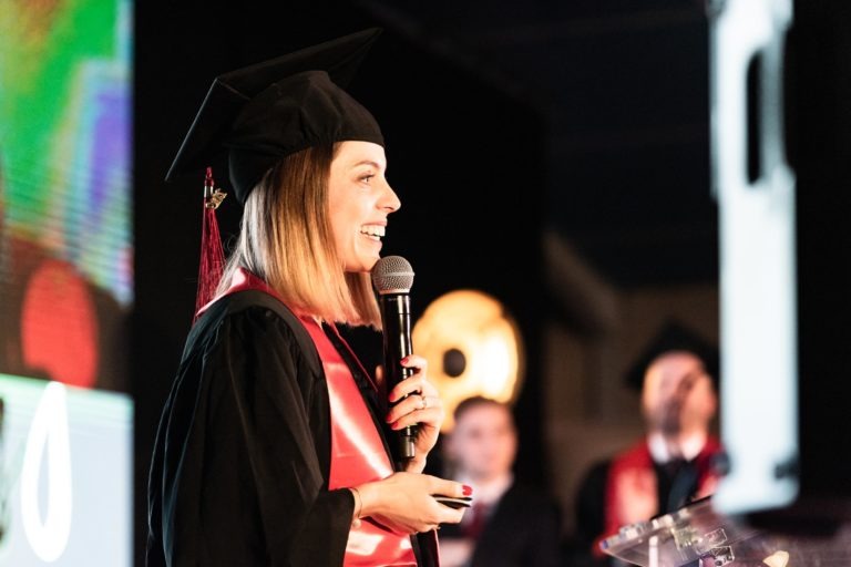 Une personne portant une toge et un chapeau de remise de diplômes, ainsi qu'une étole rouge, sourit tout en tenant un micro et en parlant sur scène lors de la cérémonie de remise des diplômes EBBS. Des silhouettes floues, également en tenue de remise de diplômes, sont visibles en arrière-plan. Le décor semble être celui d'une cérémonie de remise de diplômes en intérieur avec un éclairage lumineux et coloré.