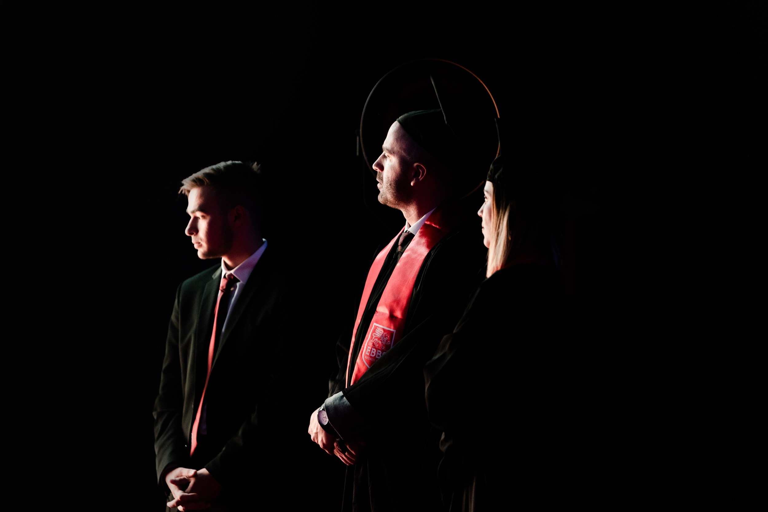 Trois personnes se tiennent debout dans la pénombre lors de la cérémonie de remise des diplômes de l'EBBS. L'homme à gauche porte un costume noir et une cravate rouge. Le deuxième homme au milieu, portant un chapeau de diplômé, une toge et une étole rouge, regarde devant lui. La femme à droite, également vêtue d'un chapeau de diplômé et d'une toge, regarde dans le vide, son profil étant faiblement éclairé.