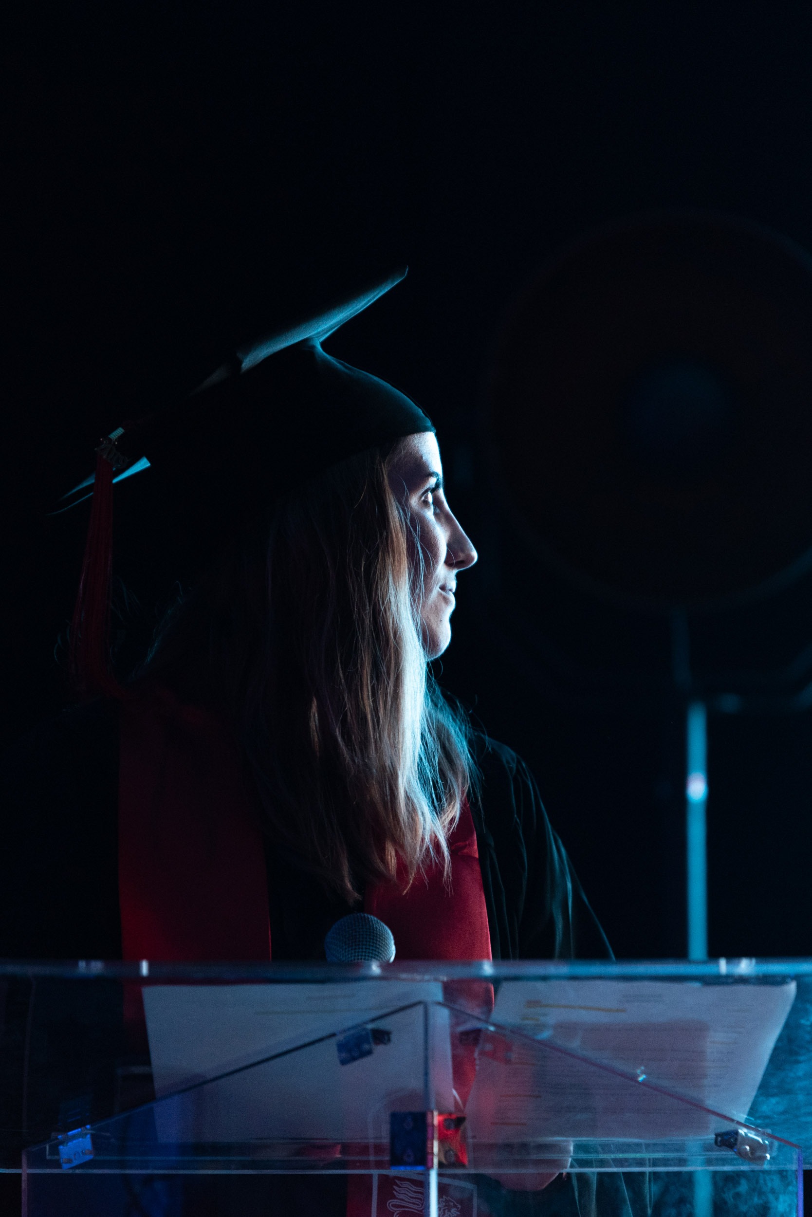 Lors de la cérémonie de remise des diplômes EBBS, une personne en toge et en toque se tient derrière un podium transparent, tenant un microphone. Le profil de l'individu est éclairé, mettant en valeur ses cheveux longs. Une étole de remise de diplôme rouge drape ses épaules. L'arrière-plan est sombre avec un luminaire circulaire partiellement visible.